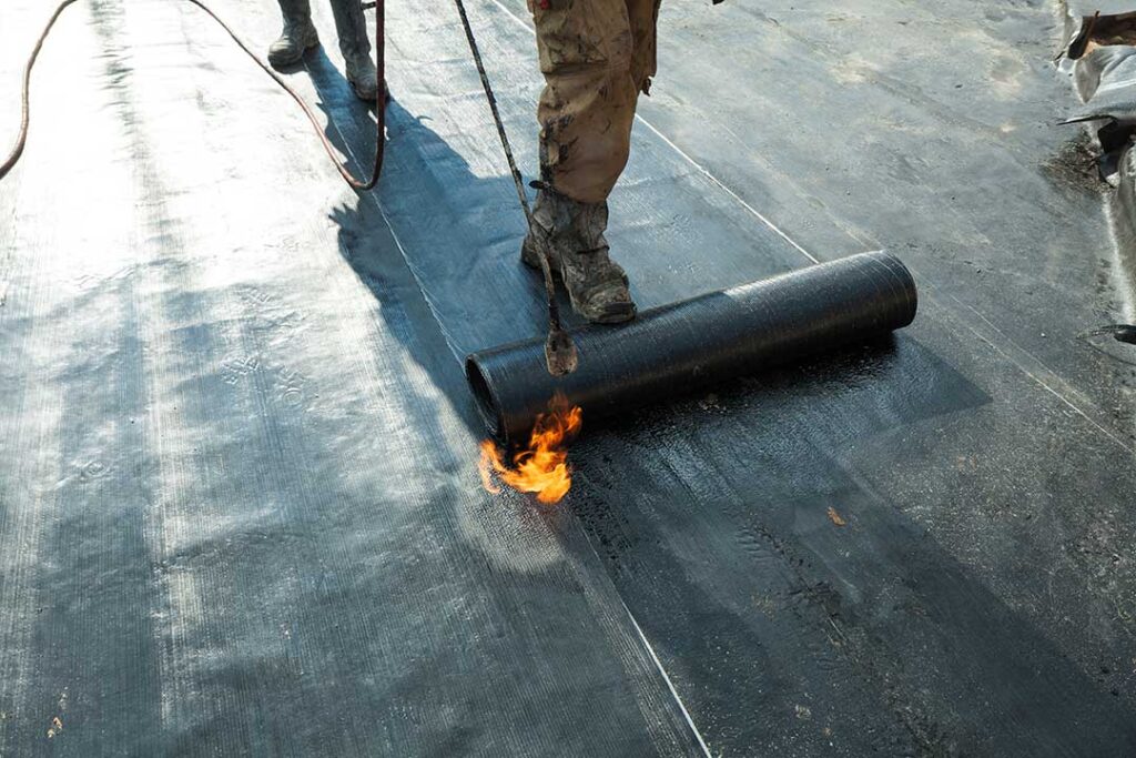 Roofing workers inspect a residential roof deck beside stacked materials and protective ground coverings.