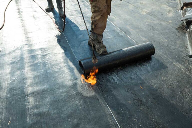 Roofing workers inspect a residential roof deck beside stacked materials and protective ground coverings.